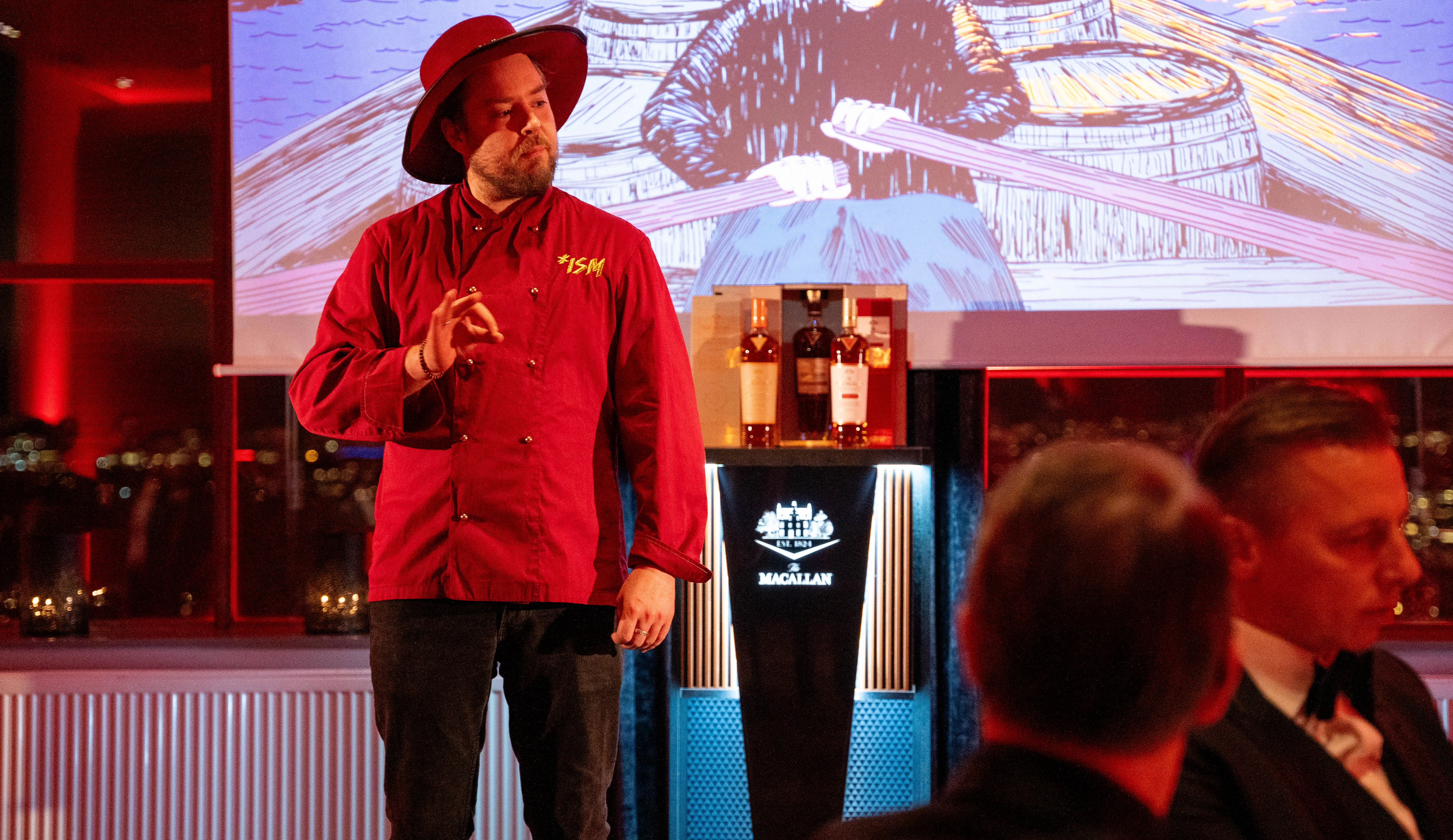 Kokk i rød jakke presenterer for gjestene, med The Macallan-montre ved siden av